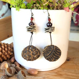 May include: A pair of dangle earrings featuring dark bronze-toned metal hooks, small red beads, dark purple spherical beads, and stacked white ceramic discs. The bottom pendants are circular, textured dark clay discs with gold-toned highlights.