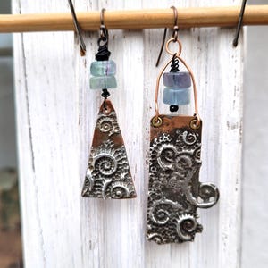 May include: Handmade earrings with a unique design. One earring is a triangle, the other is rectangular. Both have a copper-colored top and a silver-colored, textured bottom with a swirling pattern. Each earring has two blue-green beads.