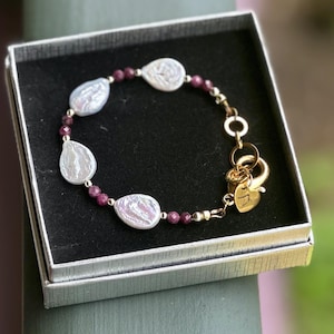 May include: A bracelet featuring white teardrop-shaped pearls and small faceted dark pink beads, connected by gold-toned metal links. The piece includes a gold-toned circular clasp and a small heart-shaped charm with engraved text inside a silver box.