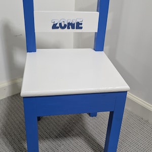 May include: A blue and white wooden chair with the words "CHILL OUT ZONE" painted on the backrest. The chair has a white seat and blue legs, perfect for a child's room or play area.