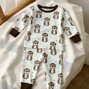 May include: White pajama set with a repeating pattern of brown cartoon otters. The long-sleeved top and pants have brown cuffs and trim. The pajamas are made of a soft, comfortable material.