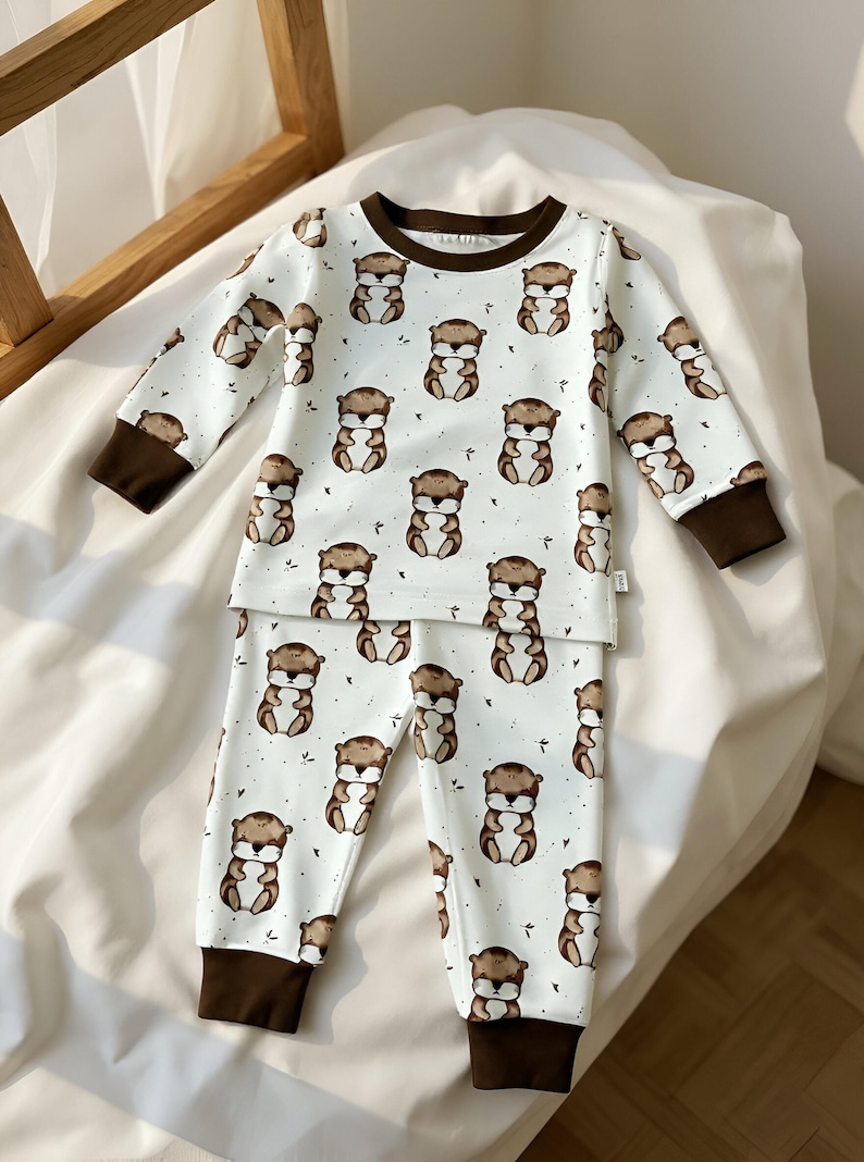 May include: White pajama set with a repeating pattern of brown cartoon otters. The long-sleeved top and pants have brown cuffs and trim. The pajamas are made of a soft, comfortable material.