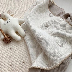 Op de afbeelding: Een cr&egrave;mekleurige babydeken met geborduurde sterren en halve manen. Een wit knuffelkonijn en een kleine rammelaar liggen naast de deken. De deken heeft een getextureerd oppervlak en een lichtbruine rand.