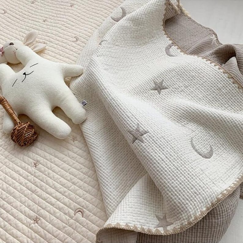 Op de afbeelding: Een cr&egrave;mekleurige babydeken met geborduurde sterren en halve manen. Een wit knuffelkonijn en een kleine rammelaar liggen naast de deken. De deken heeft een getextureerd oppervlak en een lichtbruine rand.
