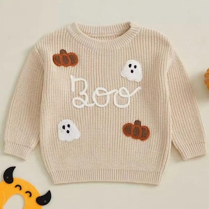 May include: A cream-coloured knitted sweater with the word "Boo" in white script. The sweater has two brown pumpkin appliqués and two white ghost appliqués. A festive Halloween-themed garment.