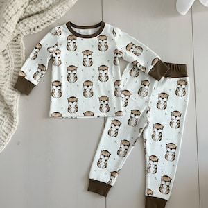 May include: White pajama set with a repeating pattern of brown otters. The long-sleeved top and pants have brown cuffs and trim. The pajamas are made of a soft material.