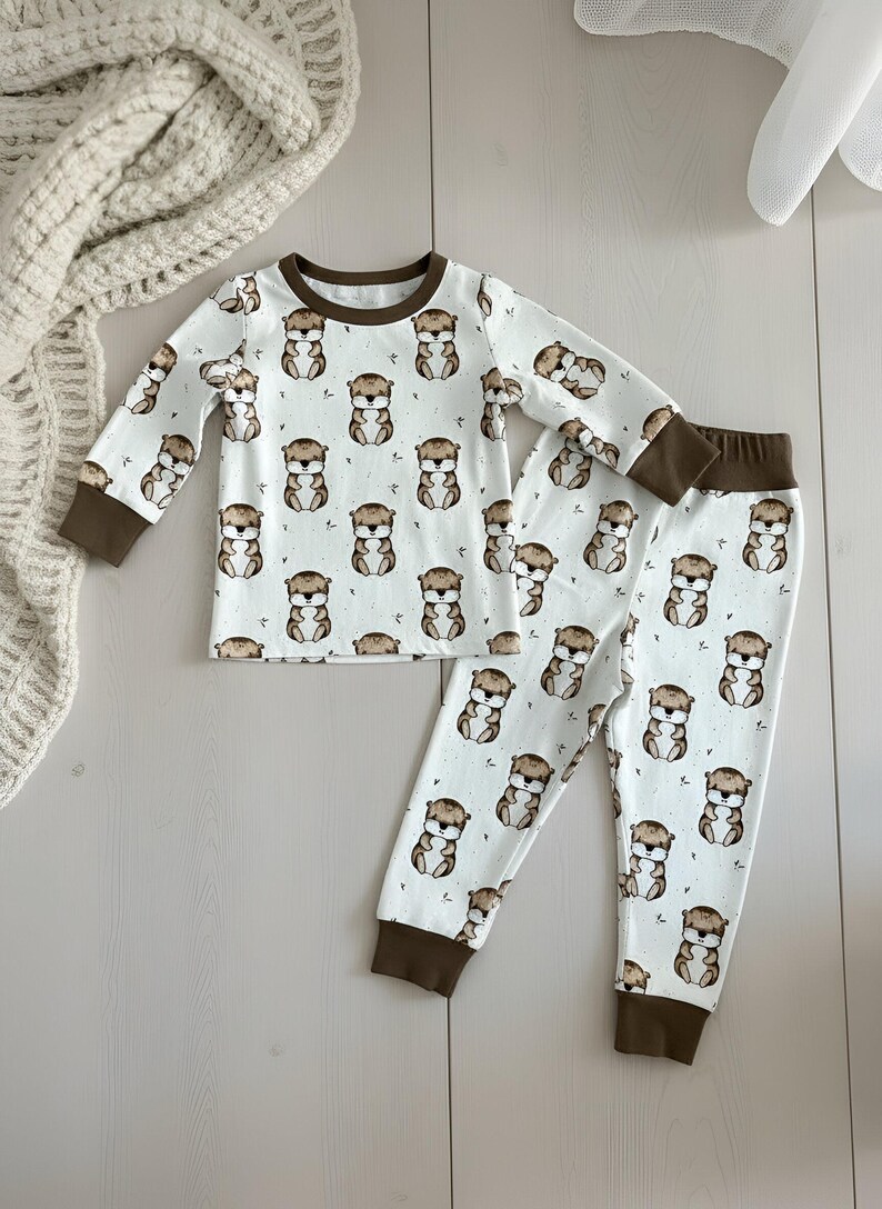 May include: White pajama set with a repeating pattern of brown otters. The long-sleeved top and pants have brown cuffs and trim. The pajamas are made of a soft material.