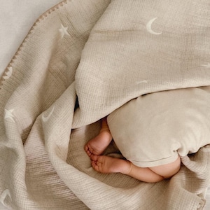 Op de afbeelding: Een beige mousseline babydeken met witte sterren en maanvormen. De deken heeft een gestikte rand en is over een baby gedrapeerd, waarvan de voeten en een deel van het lichaam zichtbaar zijn. De deken is zacht en getextureerd.