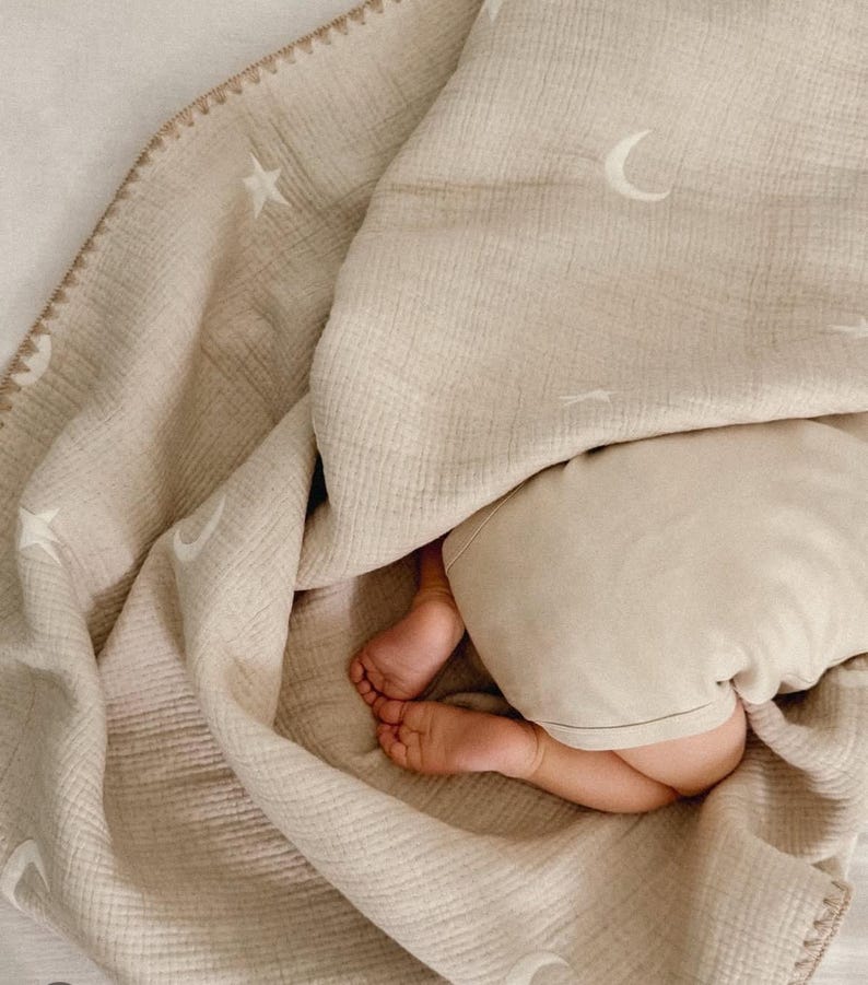Op de afbeelding: Een beige mousseline babydeken met witte sterren en maanvormen. De deken heeft een gestikte rand en is over een baby gedrapeerd, waarvan de voeten en een deel van het lichaam zichtbaar zijn. De deken is zacht en getextureerd.