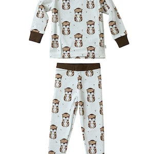 May include: A two-piece pajama set featuring a repeating pattern of brown otters on a white background. The long-sleeved top and pants have brown trim at the cuffs, neckline, and waistband. The pajamas are designed for children.