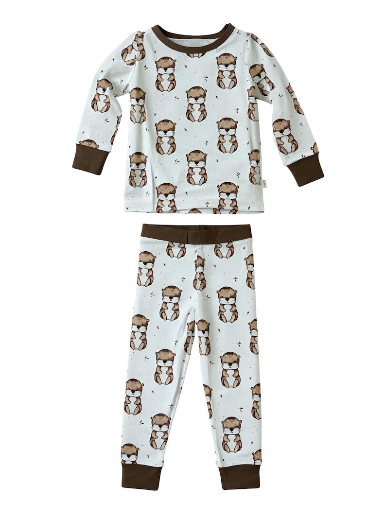 May include: A two-piece pajama set featuring a repeating pattern of brown otters on a white background. The long-sleeved top and pants have brown trim at the cuffs, neckline, and waistband. The pajamas are designed for children.