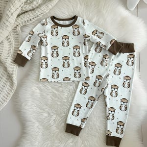 May include: A white pajama set with a repeating pattern of brown and tan otters. The long-sleeved top and pants have brown cuffs and trim. The set is displayed on a white faux fur rug.