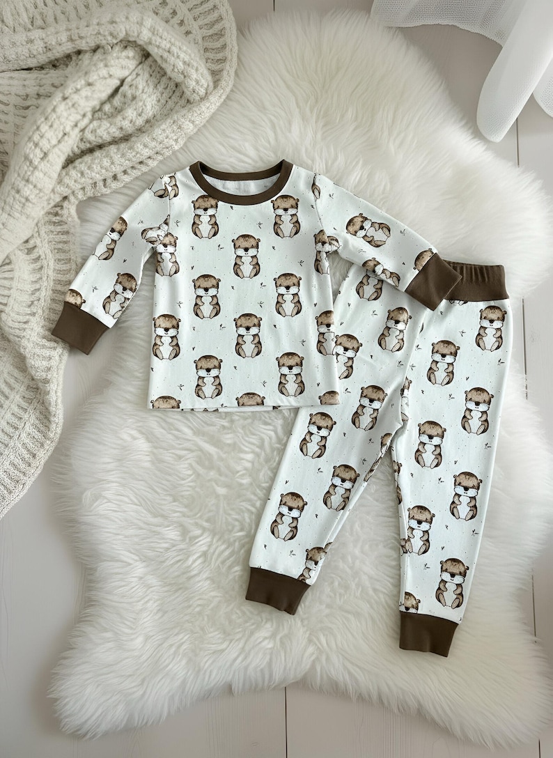 May include: A white pajama set with a repeating pattern of brown and tan otters. The long-sleeved top and pants have brown cuffs and trim. The set is displayed on a white faux fur rug.