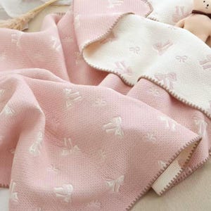 Könnte beinhalten: Eine rosa-weiße Babydecke mit Schleifenmuster. Die Decke ist wendbar, mit einer weißen und einer rosa Seite. Die Ränder sind mit einer dekorativen Naht versehen.