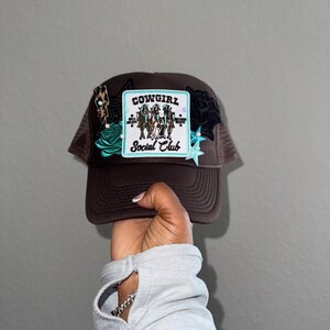 Könnte beinhalten: Eine braune Trucker-Kappe mit einem türkisfarbenen und schwarzen Patch, auf dem "Cowgirl Social Club" steht. Die Kappe hat mehrere andere Patches, darunter einen türkisfarbenen Stern, eine schwarze Rose und einen türkisfarbenen und schwarzen Blitz.