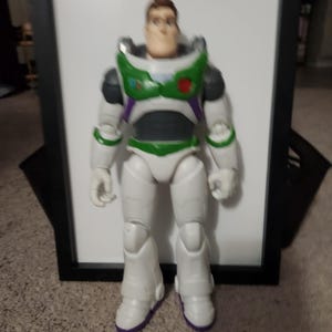 May include: A Buzz Lightyear action figure, featuring a white spacesuit with green and purple accents. The toy has a detailed face and is standing upright. The figure's boots have purple soles.