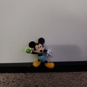 Op de afbeelding: Een Mickey Mouse-beeldje in een speelse pose, met een groene bal. Het figuur heeft zwarte oren, een geel gezicht en draagt een blauwe korte broek, gele schoenen en witte handschoenen. De achtergrond is een effen witte muur.
