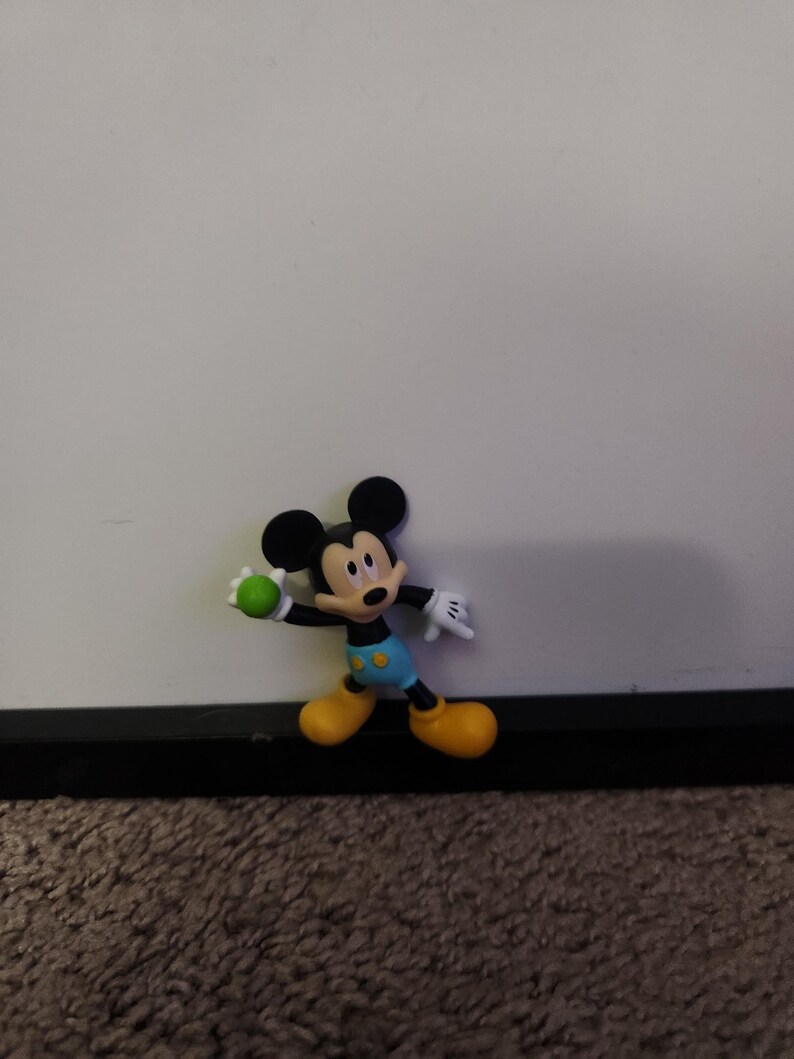 Op de afbeelding: Een Mickey Mouse-beeldje in een speelse pose, met een groene bal. Het figuur heeft zwarte oren, een geel gezicht en draagt een blauwe korte broek, gele schoenen en witte handschoenen. De achtergrond is een effen witte muur.