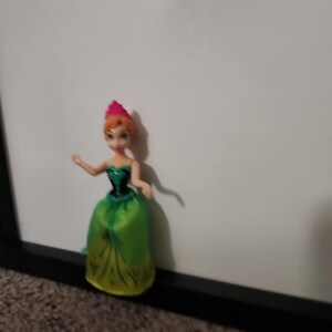 May include: A small doll of a character from the animated film Frozen. The doll has orange hair, a pink crown, and a green and yellow dress. The doll is positioned in front of a white background.