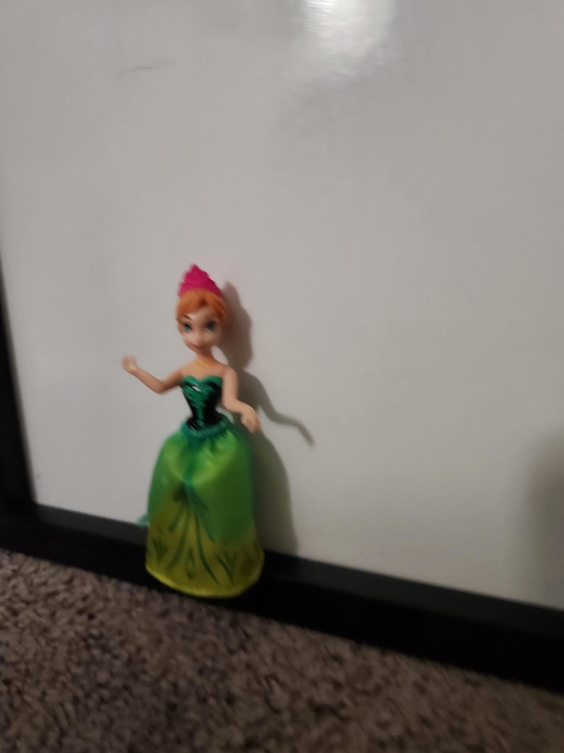 May include: A small doll of a character from the animated film Frozen. The doll has orange hair, a pink crown, and a green and yellow dress. The doll is positioned in front of a white background.