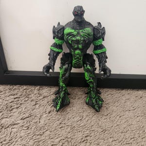 Peut inclure: Une figurine articulée noire et verte à l'aspect rocheux et texturé. Le personnage présente une carrure musclée, des mains en forme de griffes et des yeux rouges. Des reflets verts sont répartis sur le torse, les bras et les jambes.