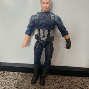 May include: A Captain America action figure in a blue and gray suit with a black belt and boots. The figure has a detailed face and is standing on a brown carpet.