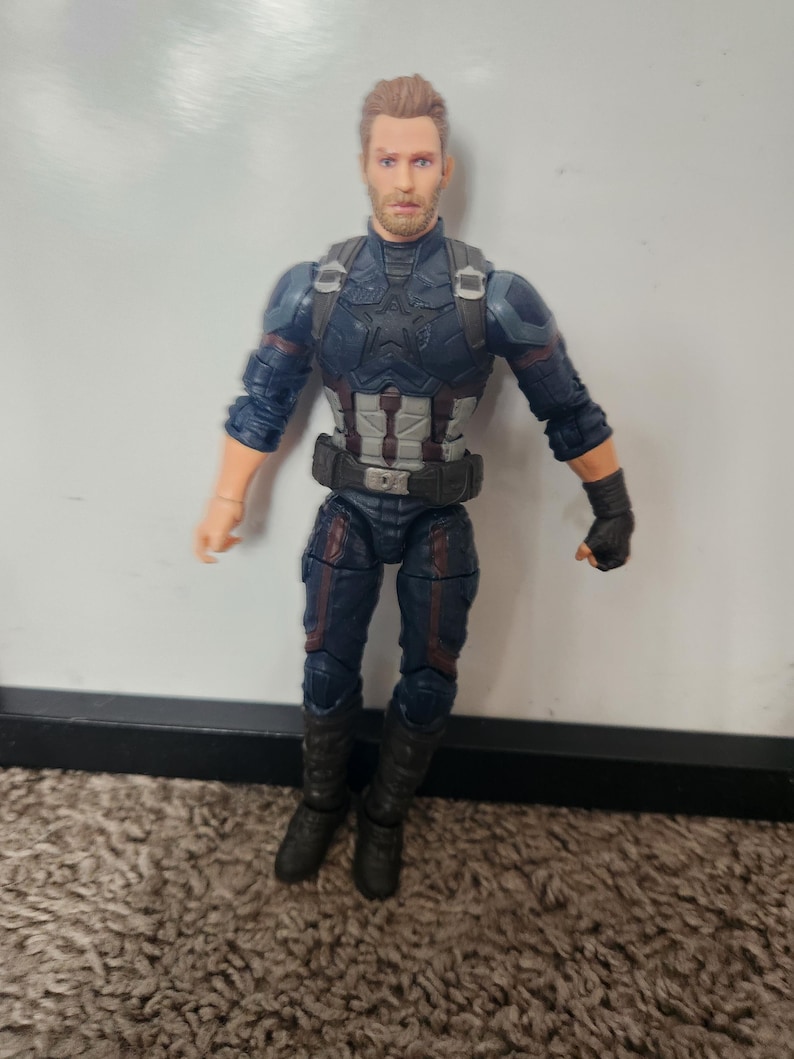 May include: A Captain America action figure in a blue and gray suit with a black belt and boots. The figure has a detailed face and is standing on a brown carpet.