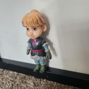 May include: A small plastic figurine of Kristoff from the movie Frozen. The figure has light brown hair, freckles, and is wearing a black and gray outfit with a red belt, green gloves, and blue boots.