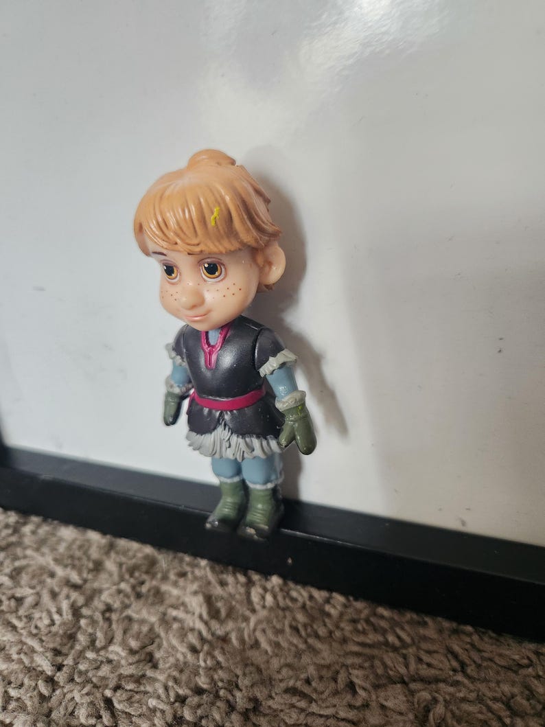 May include: A small plastic figurine of Kristoff from the movie Frozen. The figure has light brown hair, freckles, and is wearing a black and gray outfit with a red belt, green gloves, and blue boots.
