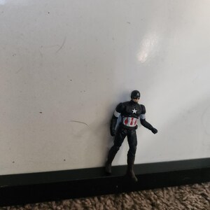 Pu&ograve; includere: Una piccola action figure di Captain America. La figura indossa un costume nero e rosso con una stella bianca sul petto e un elmetto. &Egrave; in piedi davanti a uno sfondo bianco.