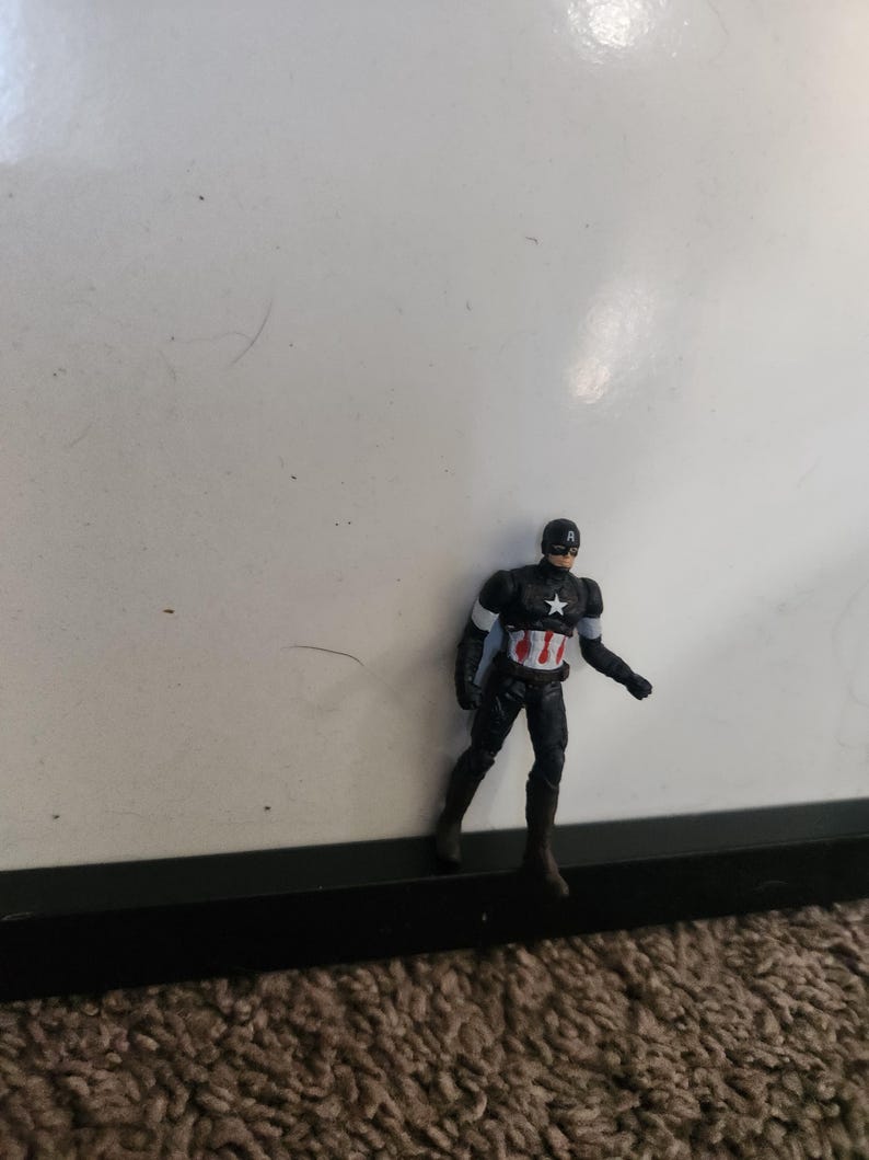 Pu&ograve; includere: Una piccola action figure di Captain America. La figura indossa un costume nero e rosso con una stella bianca sul petto e un elmetto. &Egrave; in piedi davanti a uno sfondo bianco.
