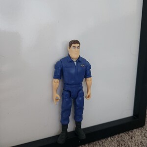 K&ouml;nnte beinhalten: Eine blaue und braune Plastik-Actionfigur eines Mannes, der einen blauen Overall mit einem Aufn&auml;her auf der linken Brust tr&auml;gt. Die Figur hat braunes Haar und steht auf einer schwarzen Oberfl&auml;che.