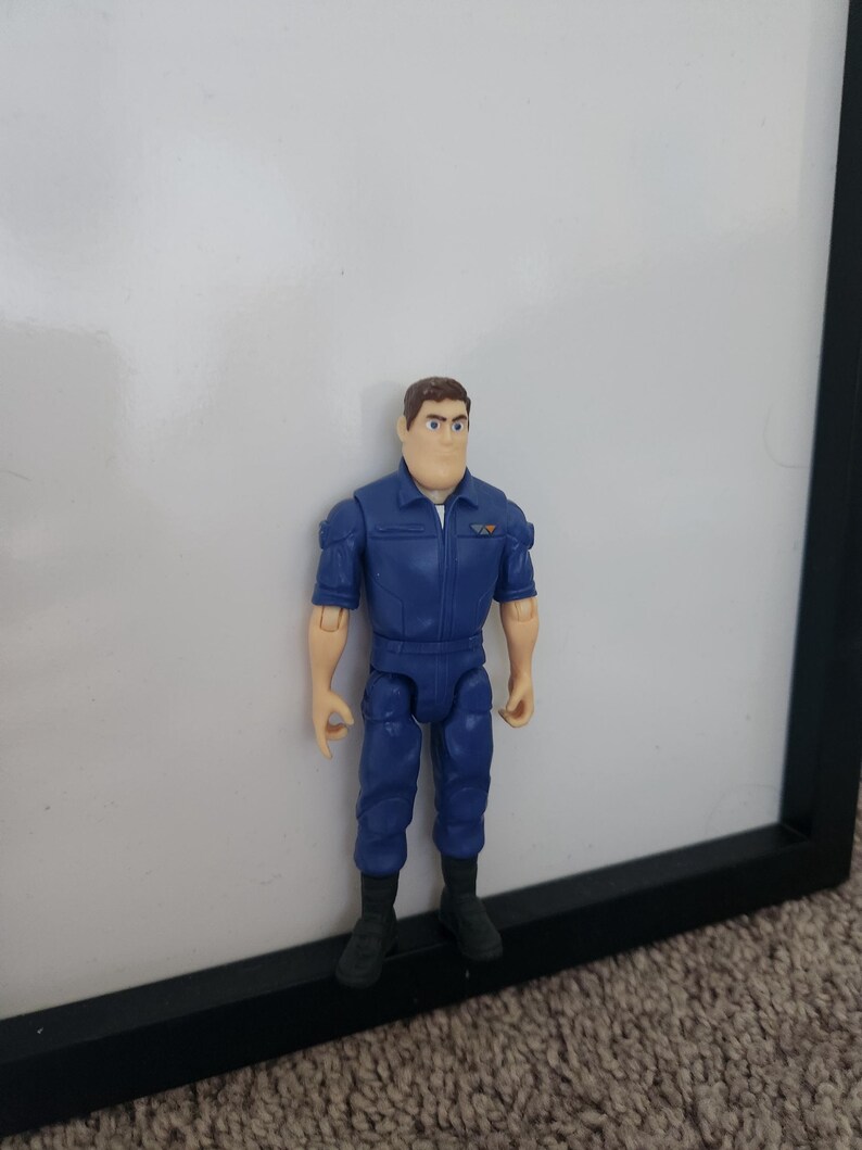 K&ouml;nnte beinhalten: Eine blaue und braune Plastik-Actionfigur eines Mannes, der einen blauen Overall mit einem Aufn&auml;her auf der linken Brust tr&auml;gt. Die Figur hat braunes Haar und steht auf einer schwarzen Oberfl&auml;che.