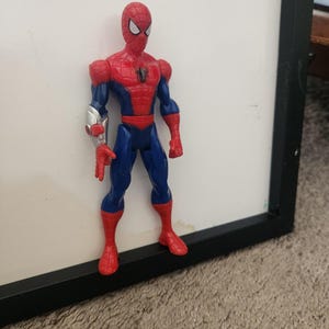 Może przedstawiać: Figurka akcji Spider-Mana w czerwono-niebieskim kostiumie z czarnym emblematem pająka na klatce piersiowej. Figurka posiada srebrny, metaliczny element na prawym przedramieniu oraz białe soczewki oczu na masce, stojąc w neutralnej pozycji.