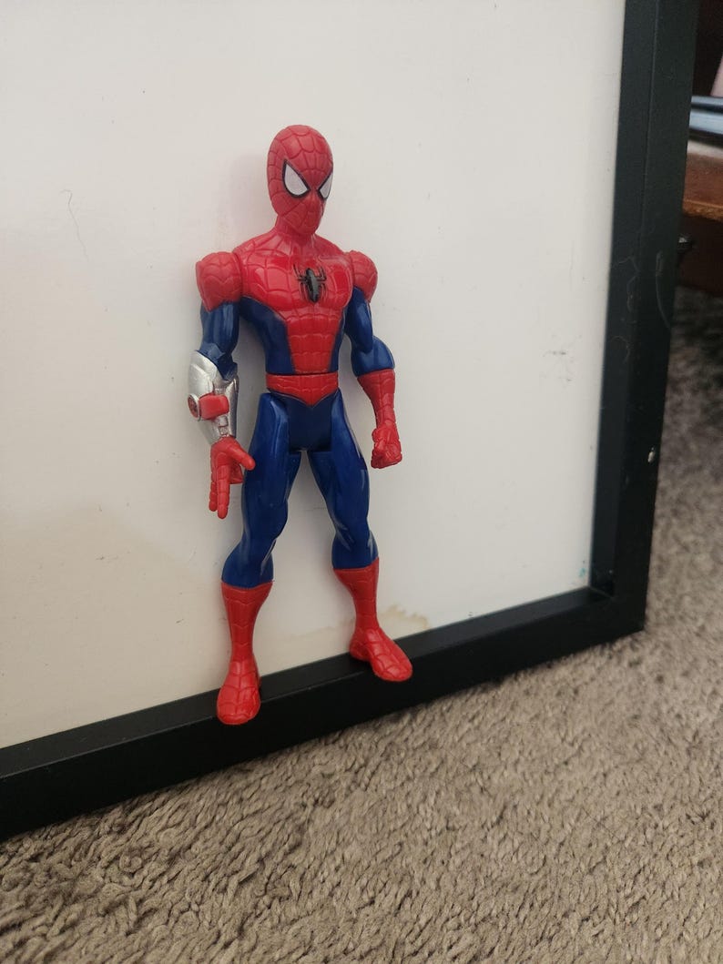 Może przedstawiać: Figurka akcji Spider-Mana w czerwono-niebieskim kostiumie z czarnym emblematem pająka na klatce piersiowej. Figurka posiada srebrny, metaliczny element na prawym przedramieniu oraz białe soczewki oczu na masce, stojąc w neutralnej pozycji.