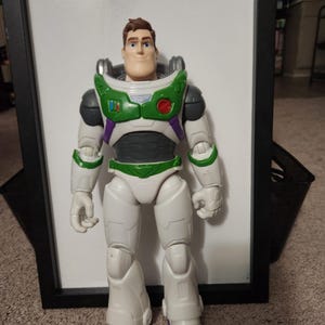 May include: A Buzz Lightyear action figure, standing upright. The figure is predominantly white with green, purple, and black accents. It features a helmet, chest armor, and boots. The figure is a toy from the Toy Story franchise.