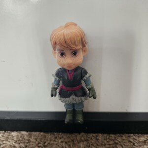 May include: A small figurine of a character with light brown hair and freckles. The figure wears a dark blue and black outfit with a red belt and green gloves and boots. The figure is standing in front of a white background.