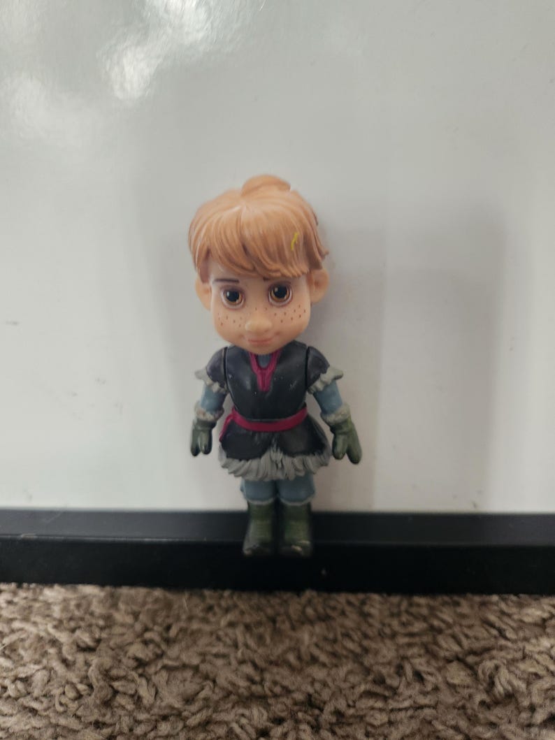 May include: A small figurine of a character with light brown hair and freckles. The figure wears a dark blue and black outfit with a red belt and green gloves and boots. The figure is standing in front of a white background.