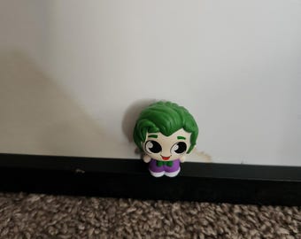 May include: A small, stylized figurine of a character with green hair, white skin, and a purple suit. The figure has large, expressive eyes and a small smile. The figurine is set against a white background and a brown carpet.