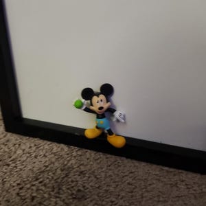 Op de afbeelding: Een kleine Mickey Mouse-figuur, met zwarte oren, een rode broek, gele schoenen, witte handschoenen en een groene bal. De figuur bevindt zich in de buurt van een zwart omkaderd wit oppervlak.