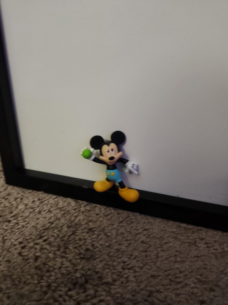 Op de afbeelding: Een kleine Mickey Mouse-figuur, met zwarte oren, een rode broek, gele schoenen, witte handschoenen en een groene bal. De figuur bevindt zich in de buurt van een zwart omkaderd wit oppervlak.