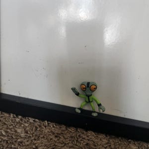 May include: A small, green alien figurine with gray skin, large orange eyes, and black accents. The figure is posed with one arm raised, standing on a black surface against a white background. The toy is a collectible.