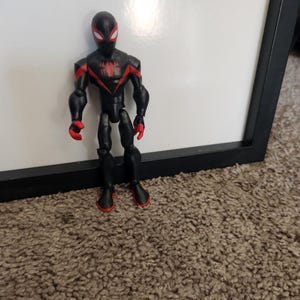 May include: A black superhero action figure with red detailing. The figure has a black mask with red eyes, a red spider emblem on the chest, and red accents on the arms and feet. It stands on a brown carpet.