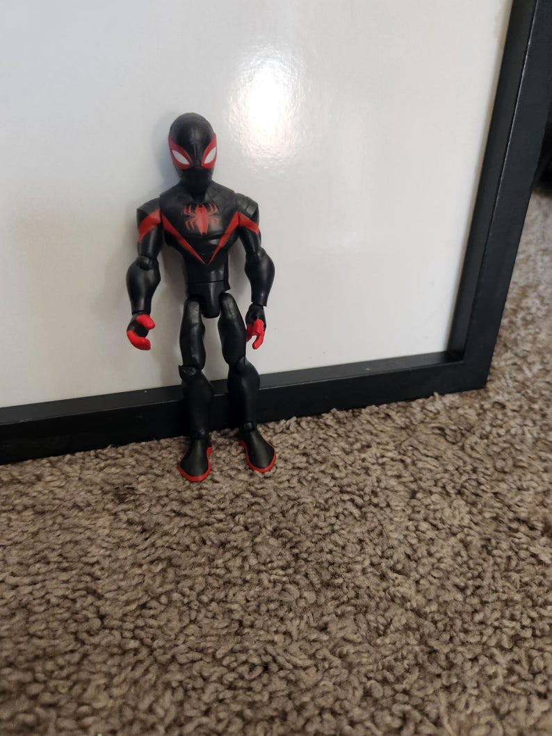 May include: A black superhero action figure with red detailing. The figure has a black mask with red eyes, a red spider emblem on the chest, and red accents on the arms and feet. It stands on a brown carpet.