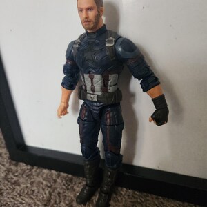 May include: A Captain America action figure in a dynamic pose. The figure is clad in a dark blue and silver suit with a utility belt and black gloves. The figure has a detailed face with a serious expression and short, styled brown hair.