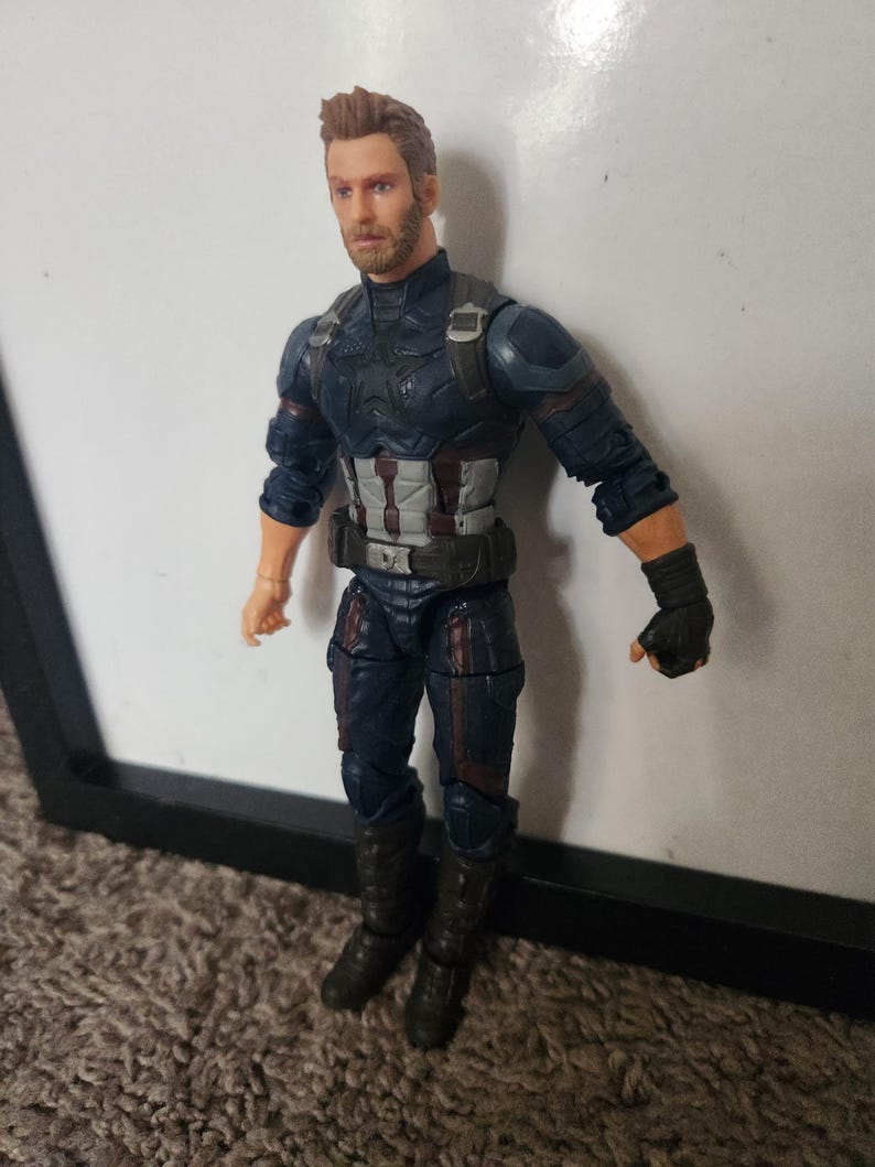May include: A Captain America action figure in a dynamic pose. The figure is clad in a dark blue and silver suit with a utility belt and black gloves. The figure has a detailed face with a serious expression and short, styled brown hair.