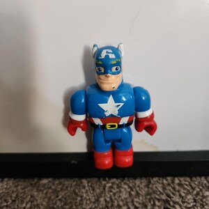May include: A blue Captain America action figure with a white star on the chest, red gloves and boots, and a black belt. The figure has a blue helmet with white wings and a painted face.