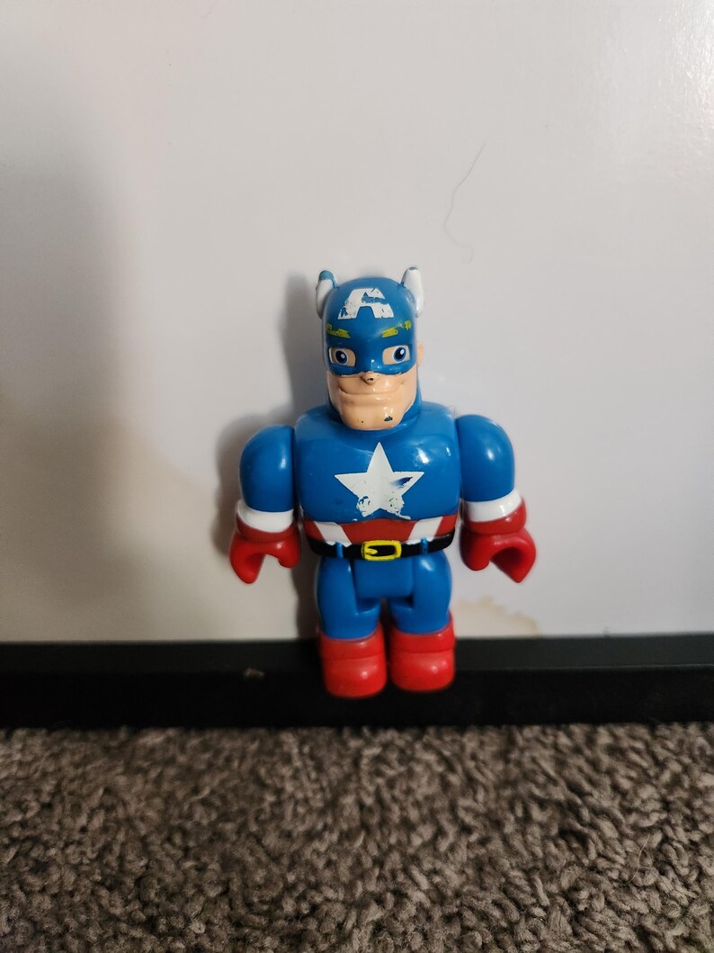 May include: A blue Captain America action figure with a white star on the chest, red gloves and boots, and a black belt. The figure has a blue helmet with white wings and a painted face.