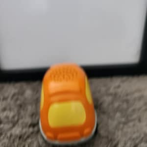 May include: A small toy car featuring a bright orange body with yellow window accents and a perforated top surface. The vehicle has a rounded, compact shape with a white base trim, showcasing a simple design with no visible wheels or interior details.