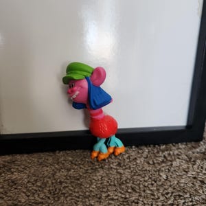 Puede incluir: Una colorida figura de muñeca Troll con piel rosa, cabello azul y un sombrero verde. La muñeca tiene un cuerpo rojo y pies turquesa y naranja. La figura está colocada frente a una superficie blanca.